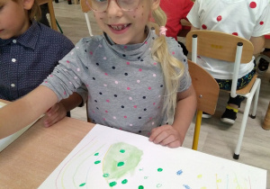 Dziewczynka z kropkami namalowanymi na twarzy i ubrana w bluzkę w kropki maluje obrazek wokół centralnie umieszczonej na nim kropki